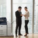 Deux personnes en tenue professionnelle dans un bureau discutent de documents à côté d'une imprimante multifonction Xerox® AltaLink® série B8200